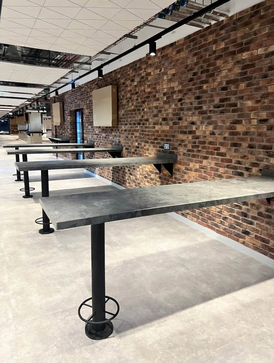Zinc-topped table pedestals with powder coated bases along exposed brick wall at Everton Stadium Village Street